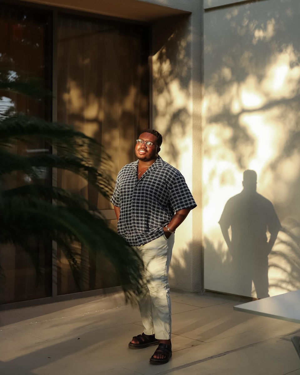 Young man standing outdoors with sunlight and shadows on wall, shadow of person visible, wearing glasses, patterned shirt, light pants, sandals