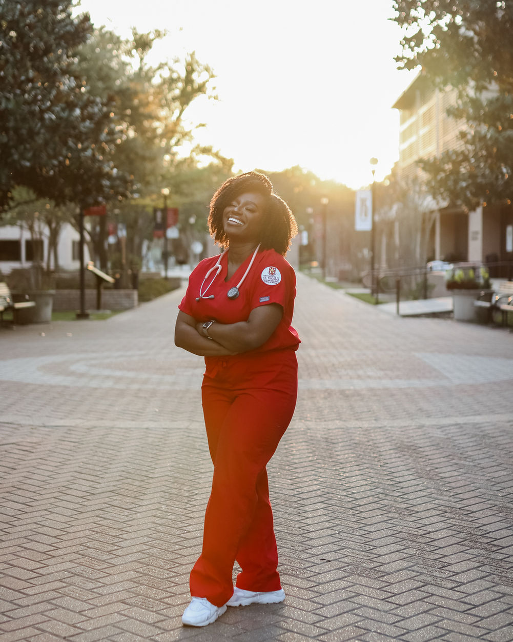 Person in red medical scrubs standing outdoors on a paved walkway, smiling with arms crossed, trees and buildings in background