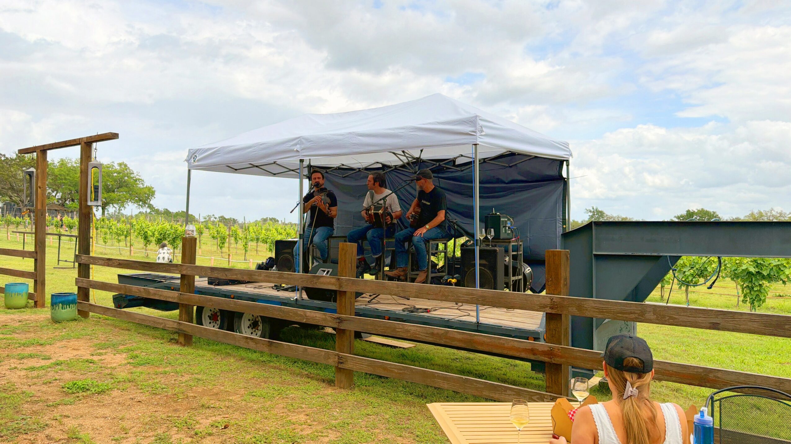 People performing music on a stage under a canopy outdoors, with a wooden fence and grassy field nearby.