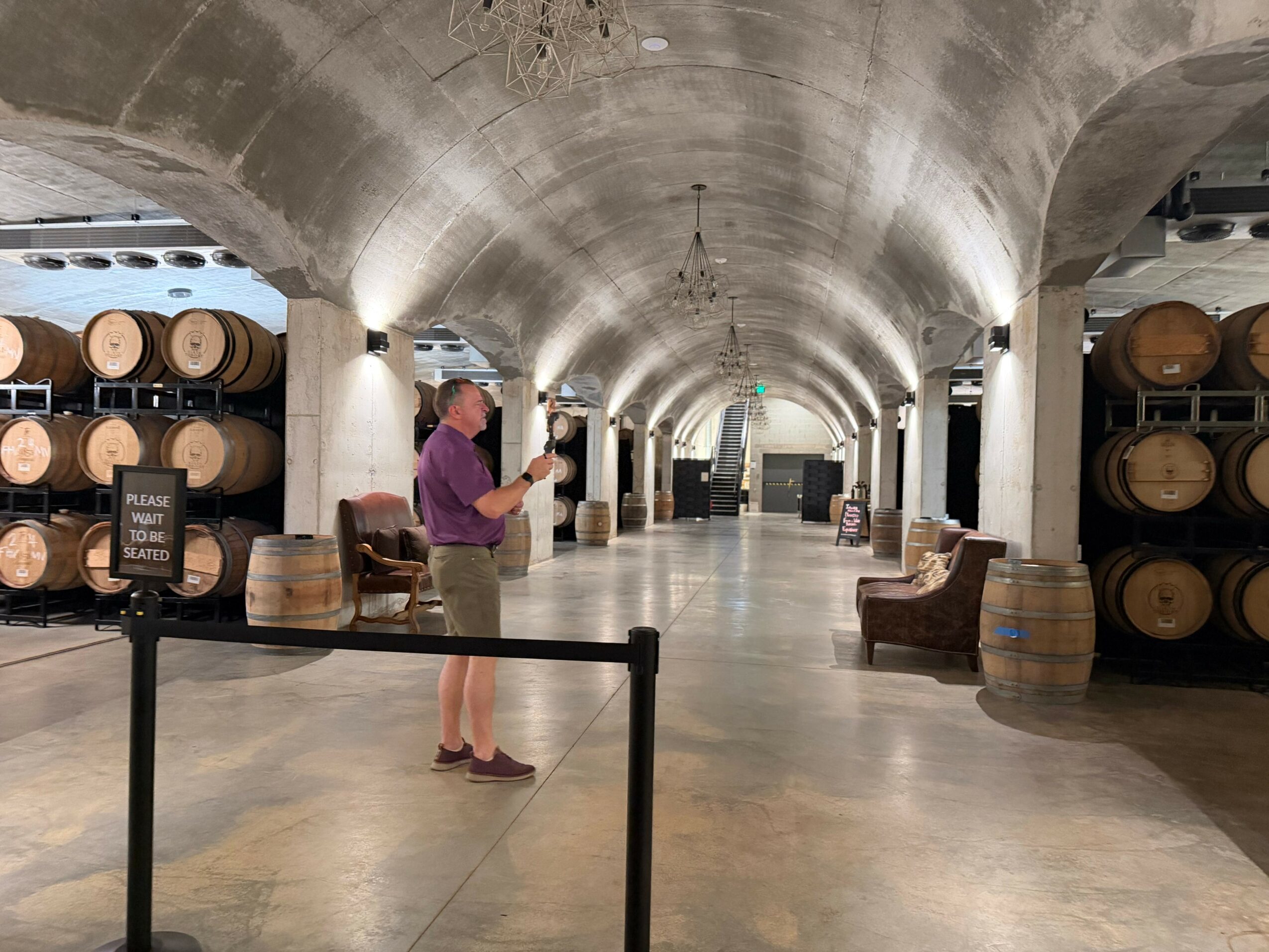 Person in purple shirt and shorts reading a menu in a wine cellar with barrels and arched ceiling.