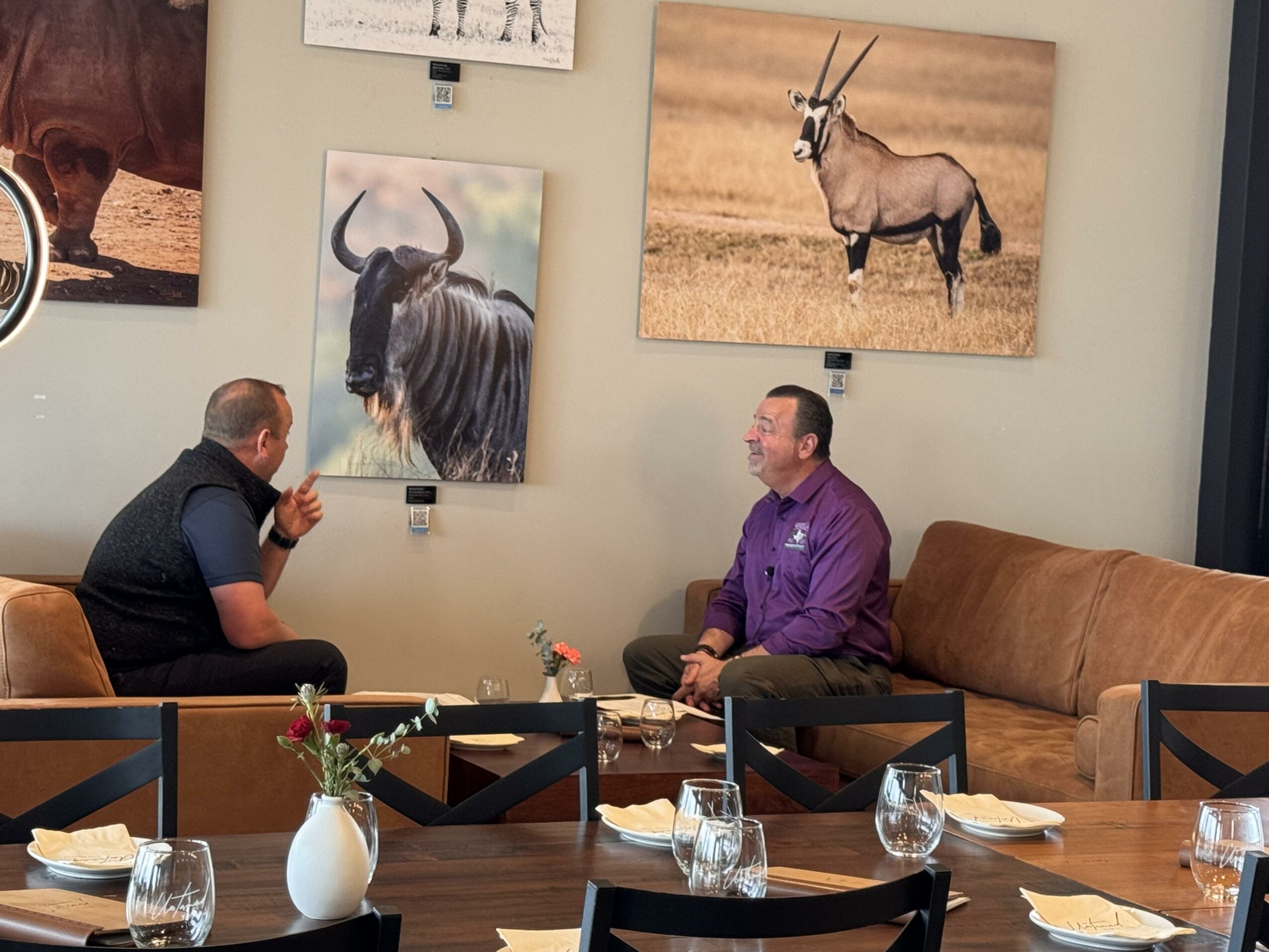 Two men sit on a brown couch and talk, with a table and chairs in front, and animal photographs on the wall behind.