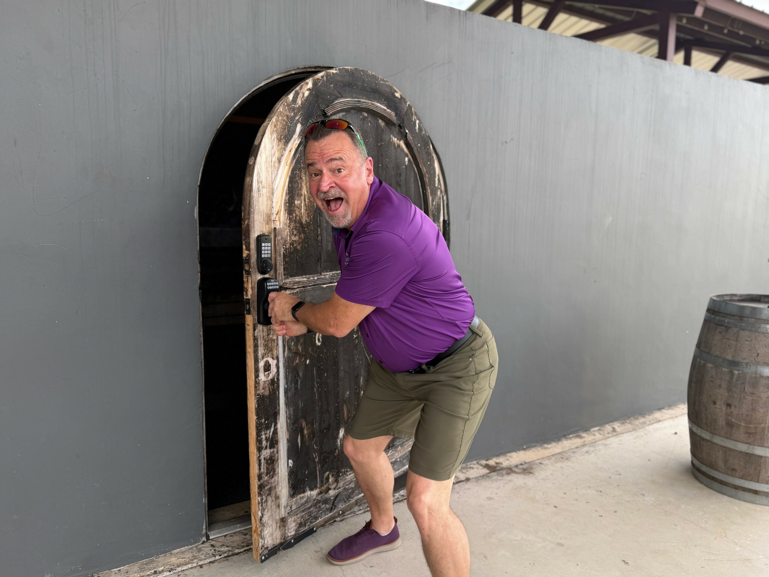 Man in purple shirt and shorts opening a small door in a gray wall, with a barrel nearby.