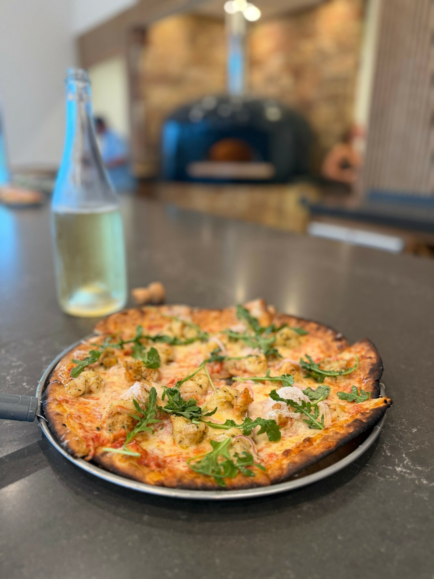 Cheese pizza with herbs on a black plate, bottle of beverage in background, in a restaurant setting.