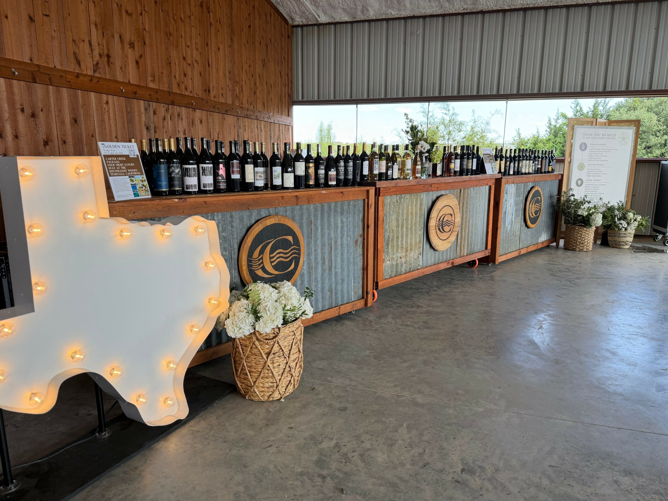 Bar area with bottles on a wooden counter, a large illuminated Texas-shaped sign with lights, and a basket of white flowers.