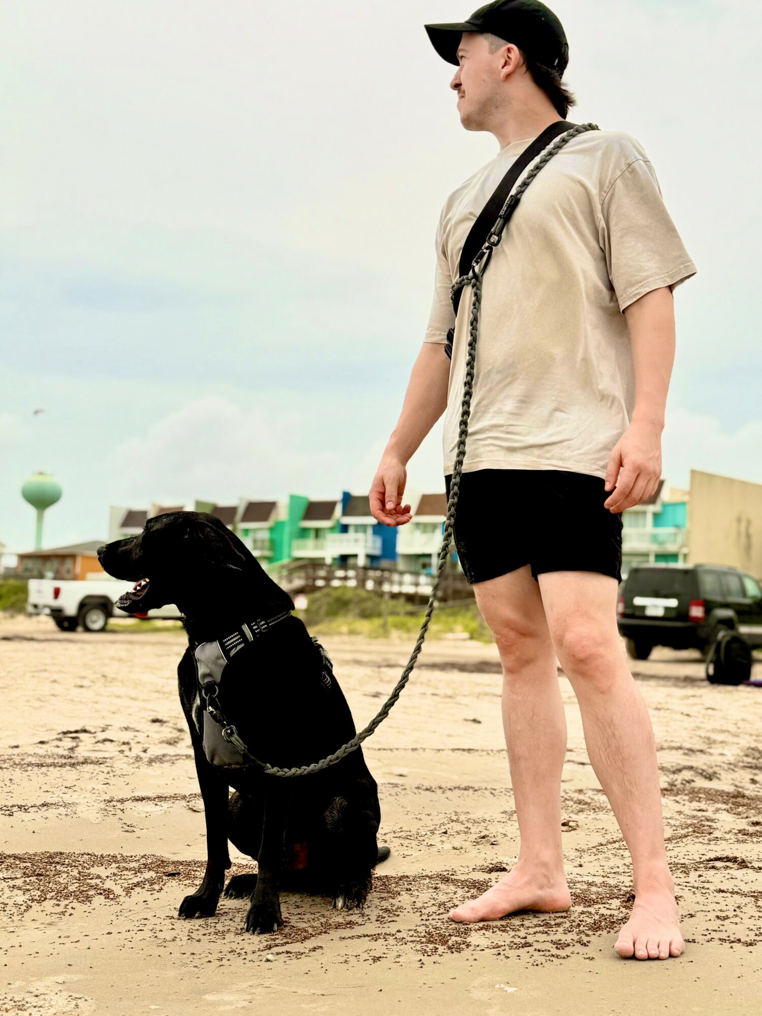 Person standing barefoot on sandy ground with a black dog sitting beside them, holding a leash, outdoors with buildings and vehicles in background.