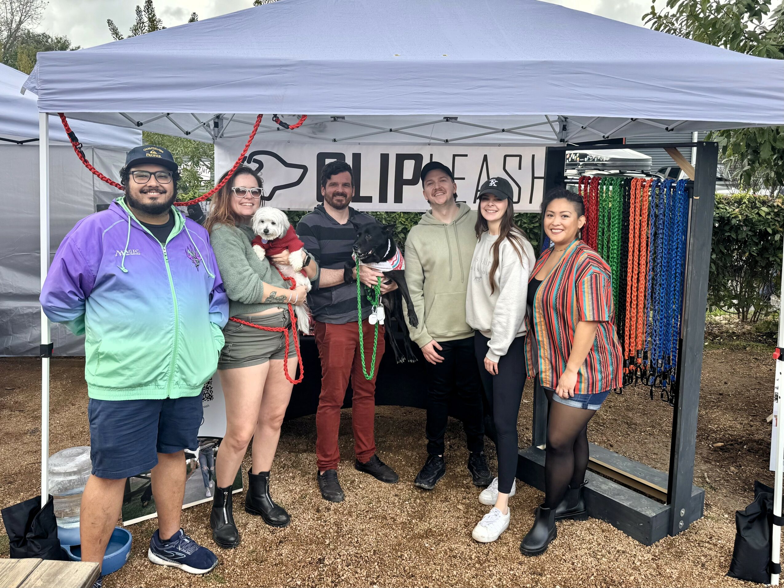 Group of six people standing under a white canopy with dog accessories and colorful leashes displayed behind them.
