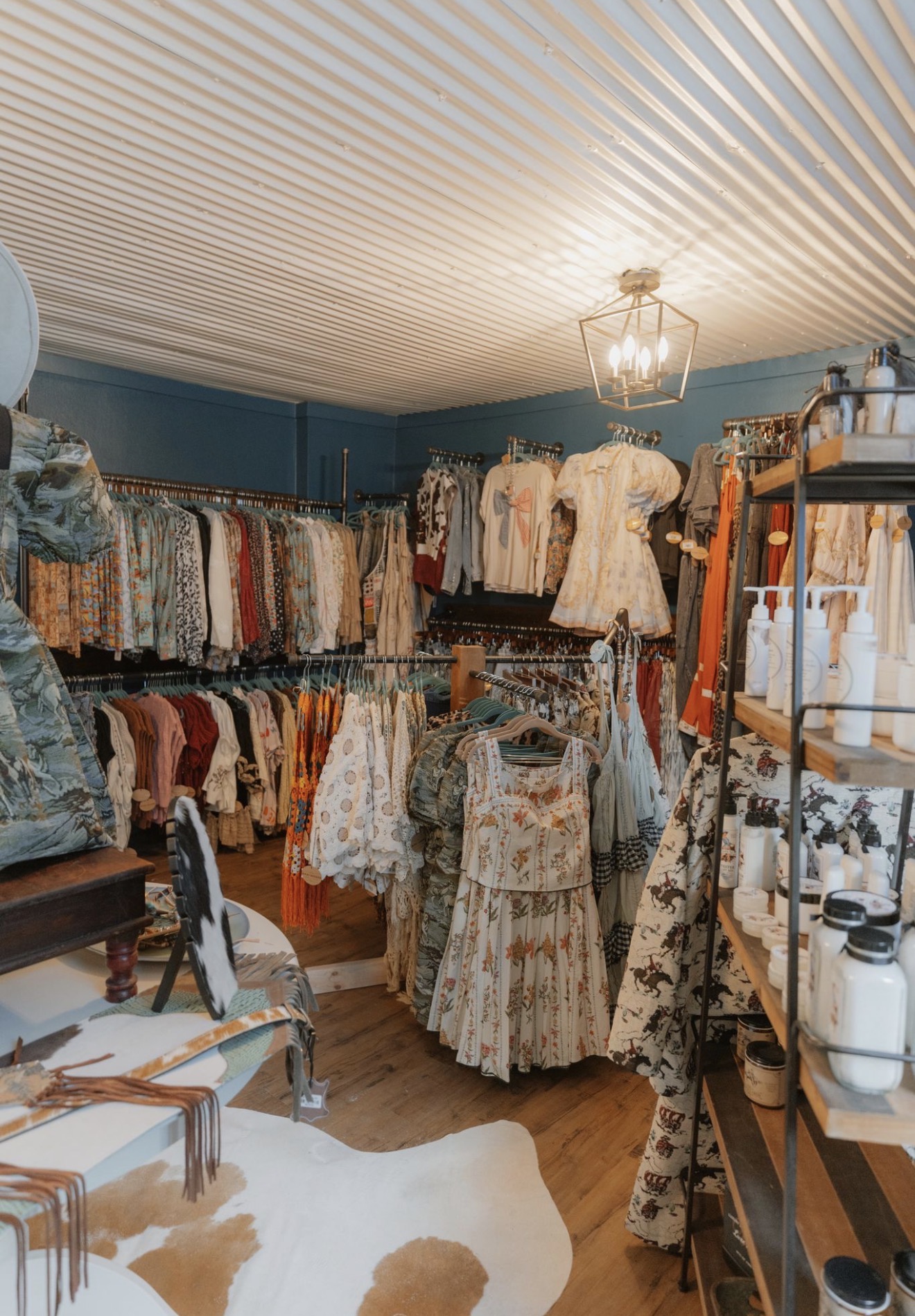 Clothing store interior with racks of dresses, sweaters, and accessories, wooden shelves, and a ceiling light fixture.