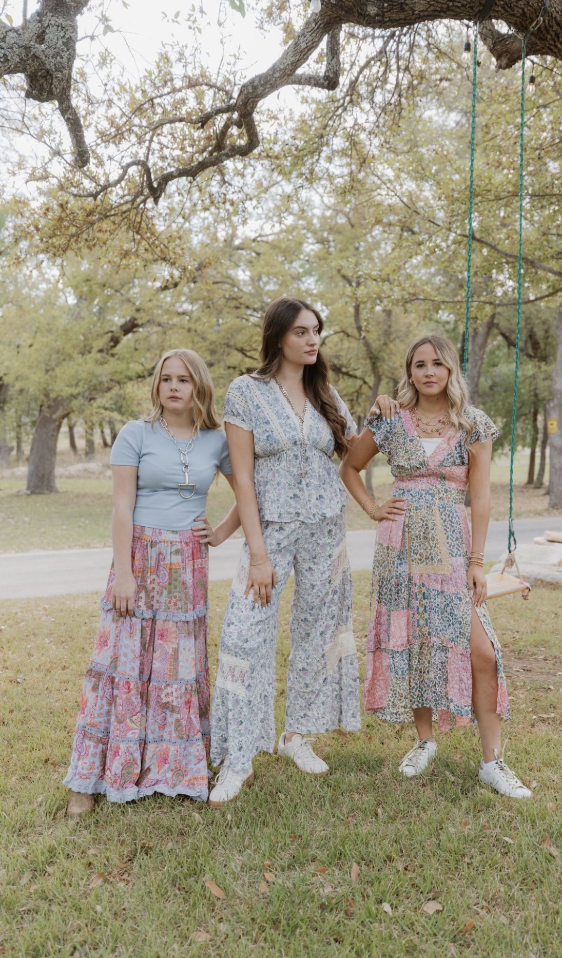 Three women standing outdoors under a tree, wearing colorful patterned outfits, with a swing in the background.