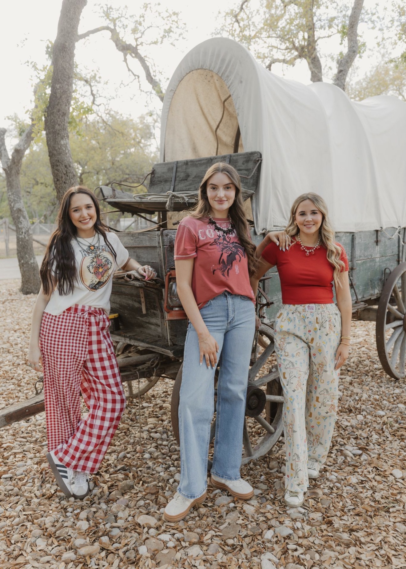Three women stand outdoors near a covered wagon, surrounded by trees and fallen leaves, smiling at the camera.