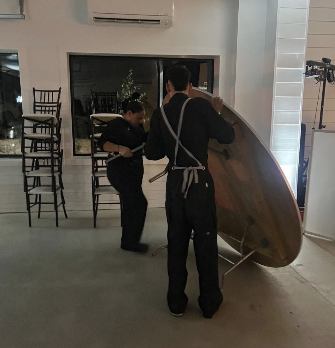 Two people setting up a round table indoors, with stacked chairs and a window in the background.