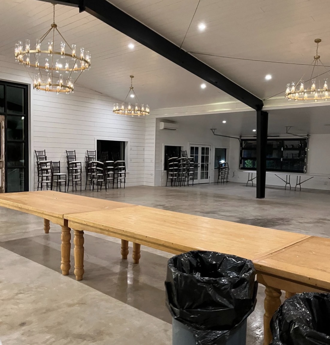 Long wooden table in a spacious room with chandeliers, chairs, and a black trash bag on the floor.