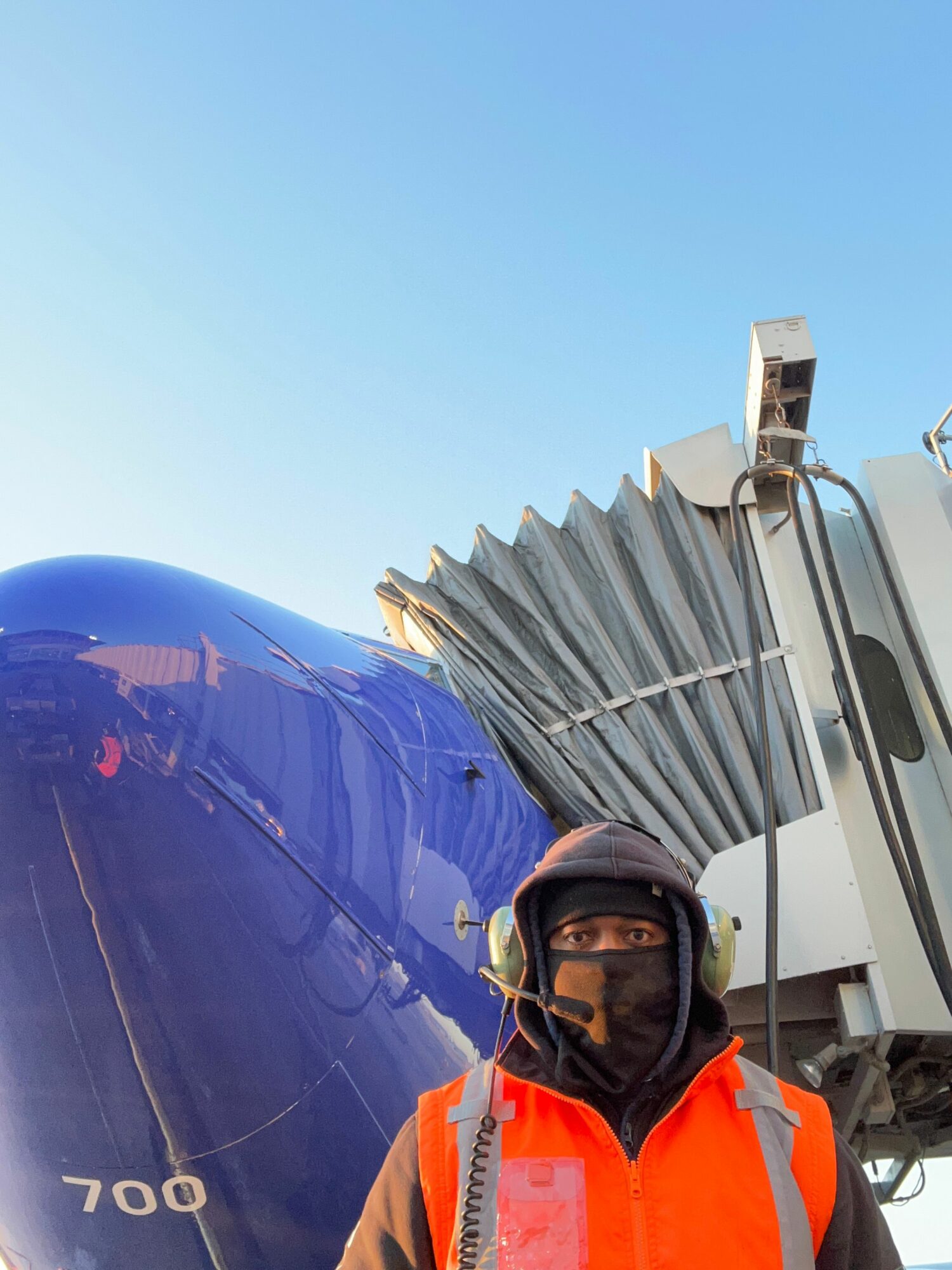 Person wearing safety gear stands next to a large blue object with a clear sky background.