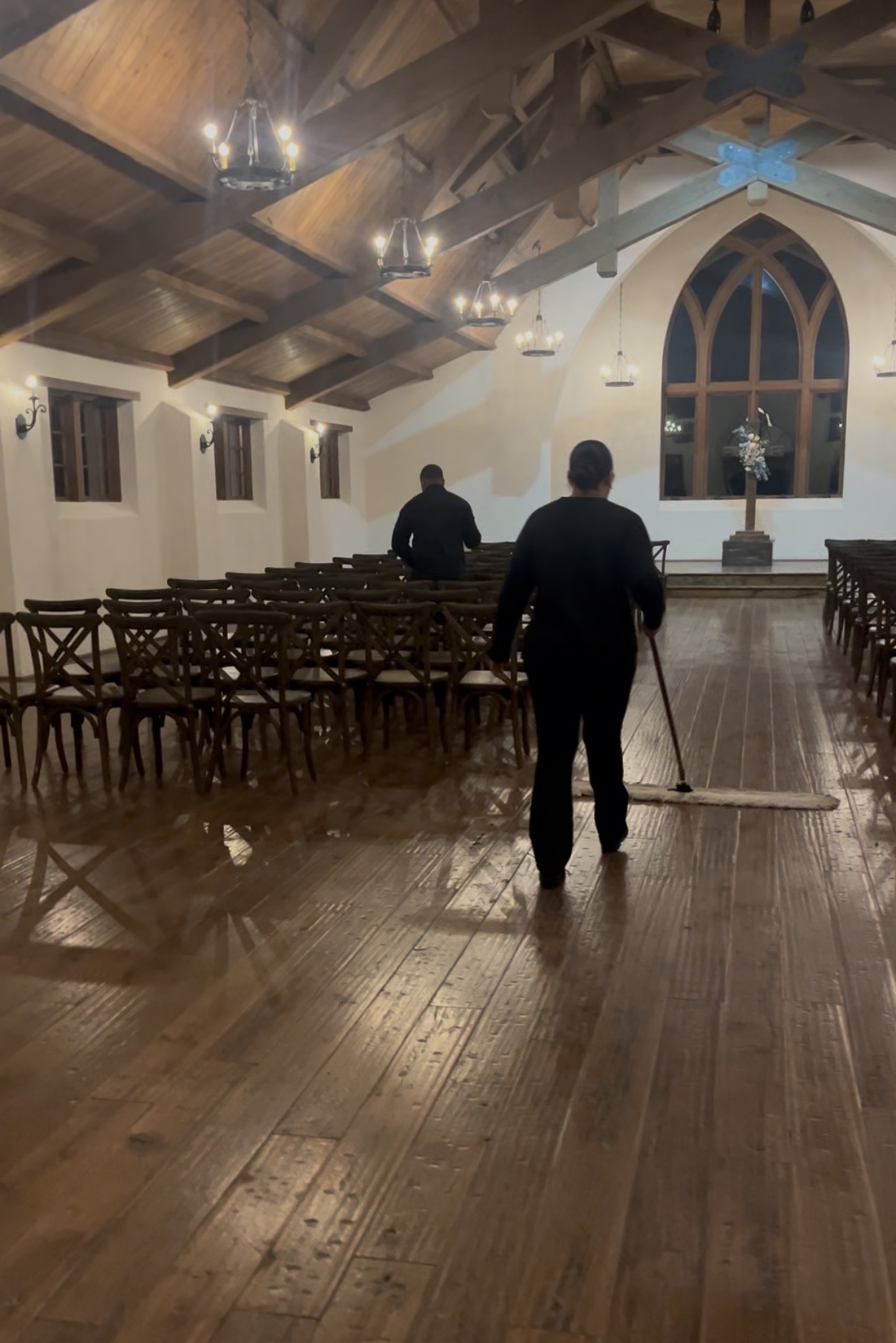 Two people cleaning a wooden floor inside a church with wooden chairs and a large arched window, ceiling with exposed beams.