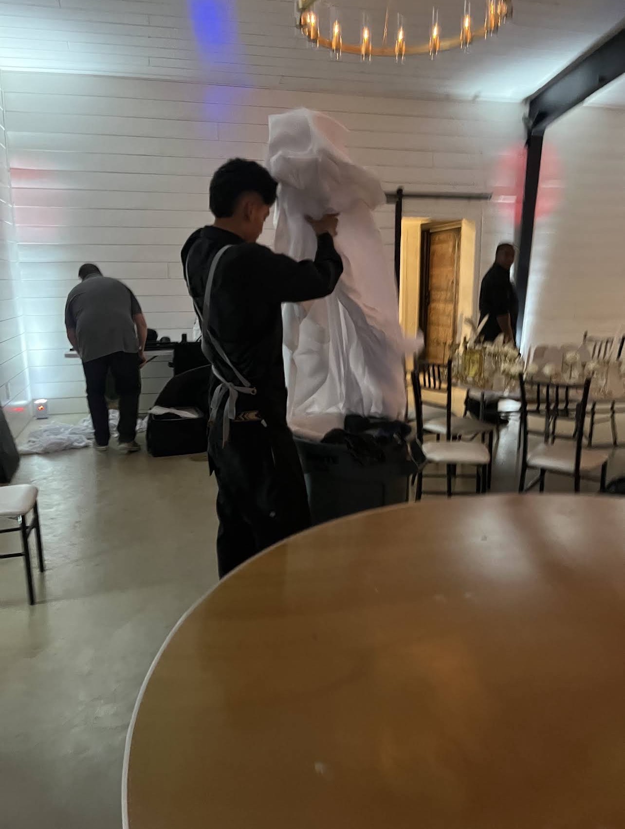 Person adjusting a large white fabric or costume in a decorated indoor space with chairs and other people present.