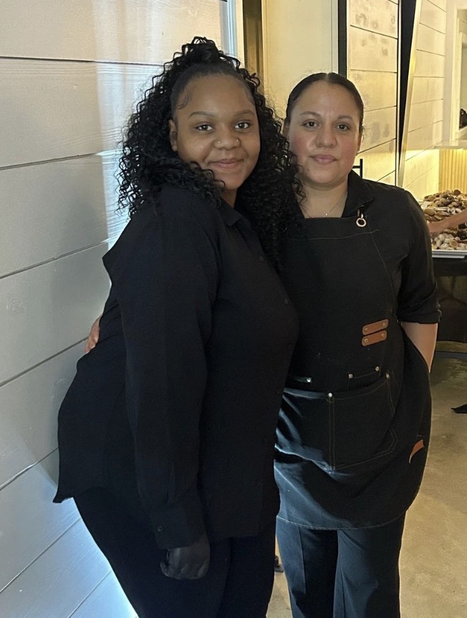 Two women standing indoors, smiling, one with curly hair and the other with straight hair, wearing black clothing.