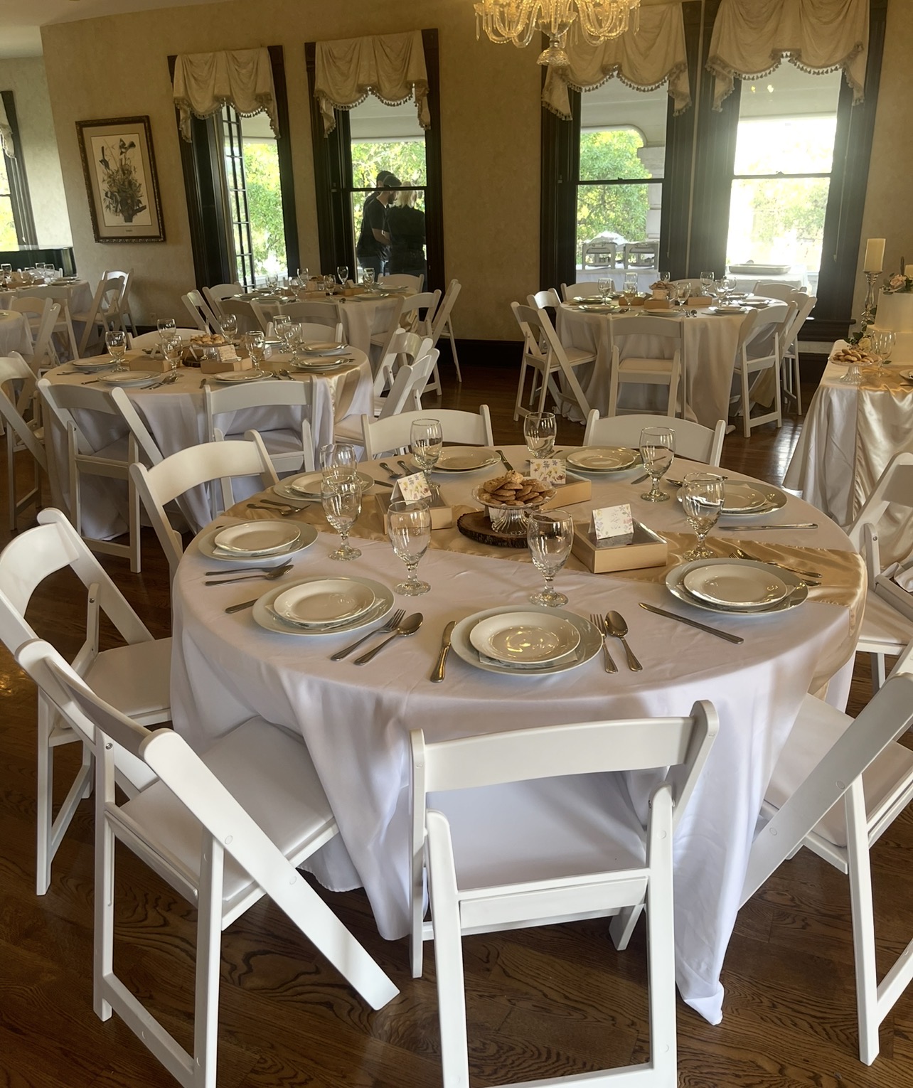 Round table set with plates, glasses, and silverware in a decorated dining room with large windows and natural light.