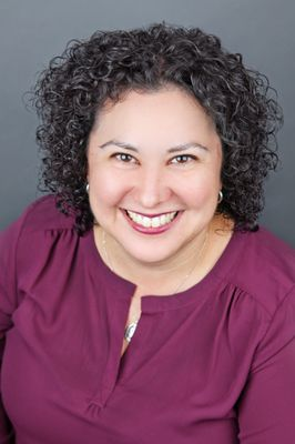 Woman with curly hair smiling, wearing a purple top, against a gray background.