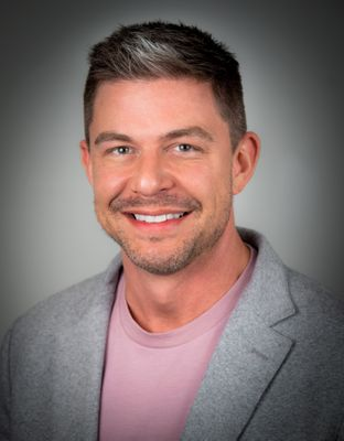 Portrait of a smiling man with short dark hair, wearing a light pink shirt and gray blazer, against a gray background.
