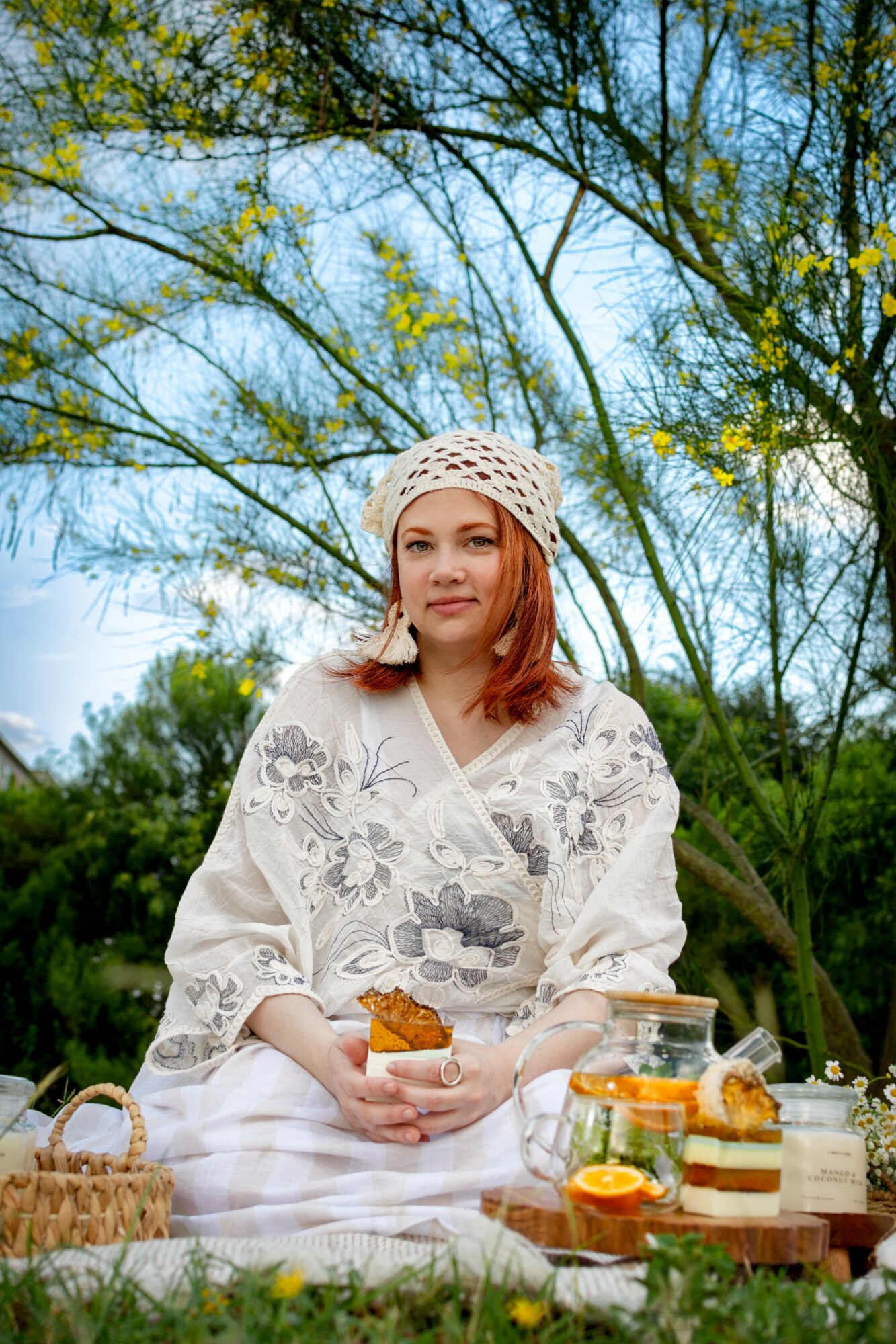 Young woman with red hair sitting outdoors, holding a cup, surrounded by food and drinks, trees in background.