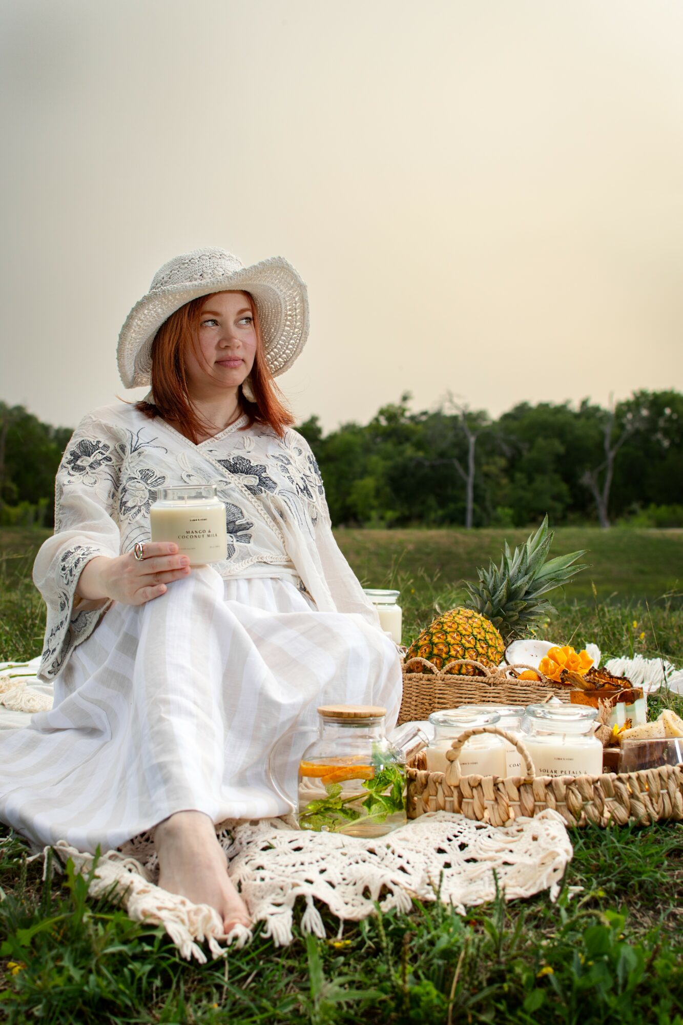 Woman in a white dress and hat sitting outdoors with a picnic spread, holding a drink, surrounded by greenery.