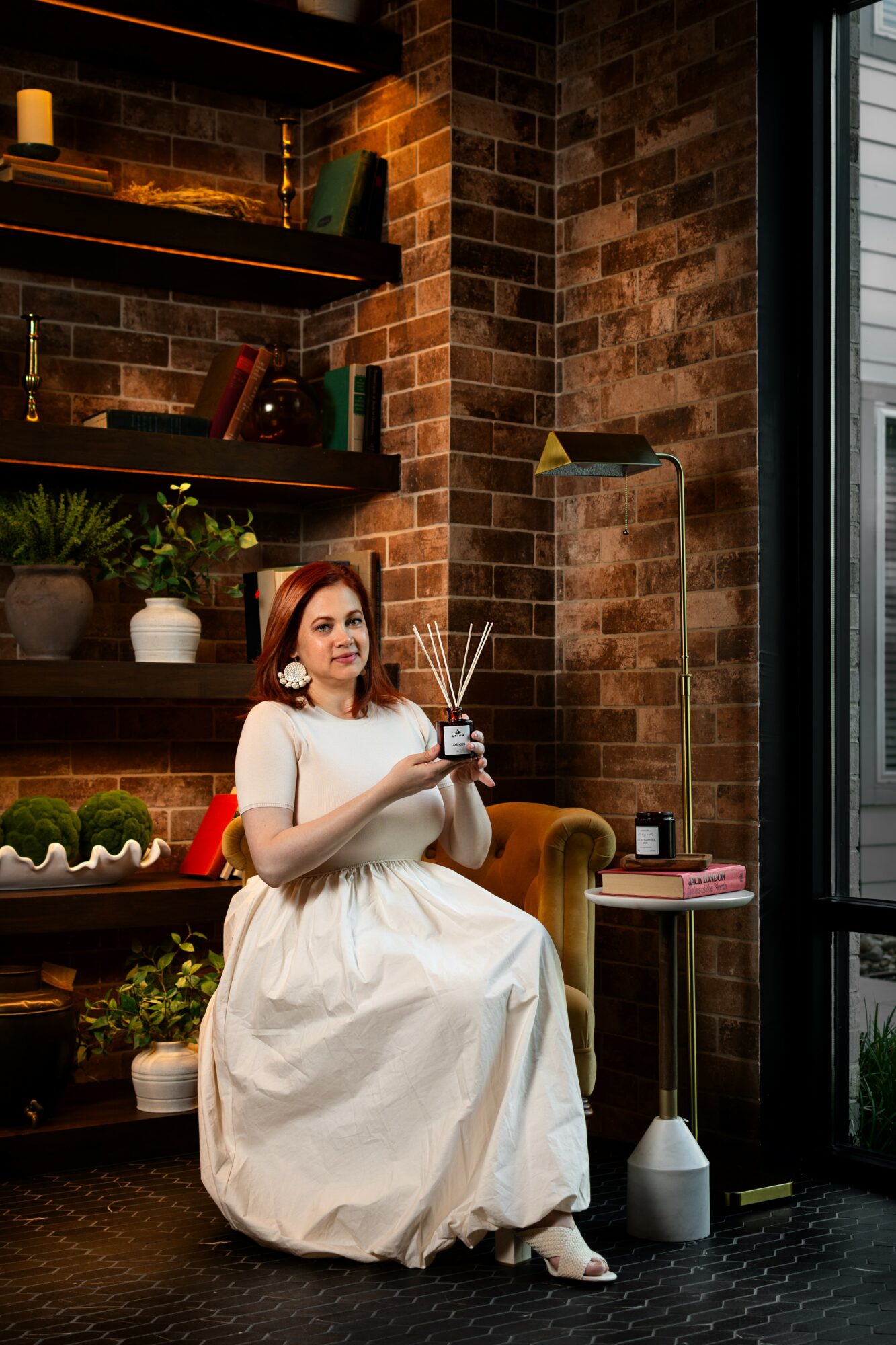 Woman in white dress sitting on a yellow armchair holding a cup and a spoon, in a brick-walled room with plants and shelves.