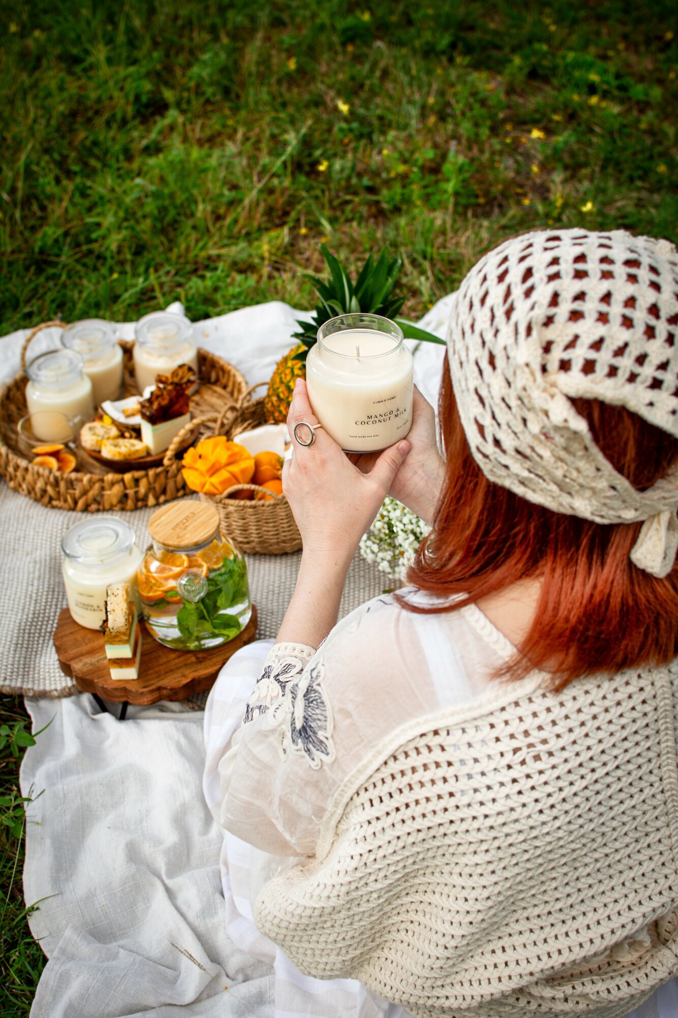 Person with red hair and white hat holding a glass jar at outdoor picnic with food and drinks on a blanket.