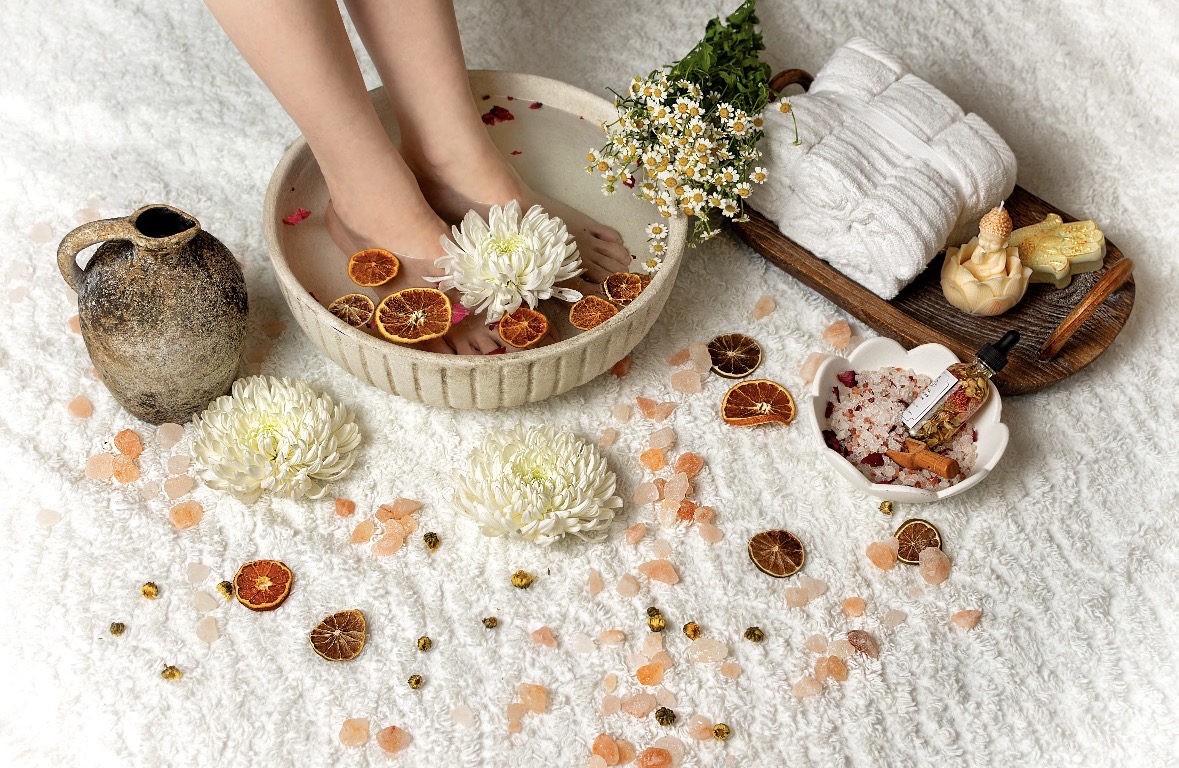 Person soaking feet in a bowl with flowers, surrounded by towels, flowers, and spa items on a white surface.