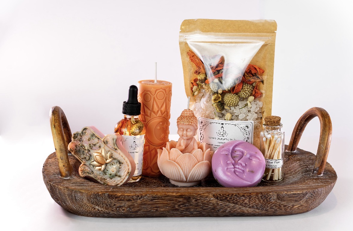 Wooden tray holding a candle, dropper bottle, Buddha statue, purple soap, and packaged herbs or tea, against a white background.