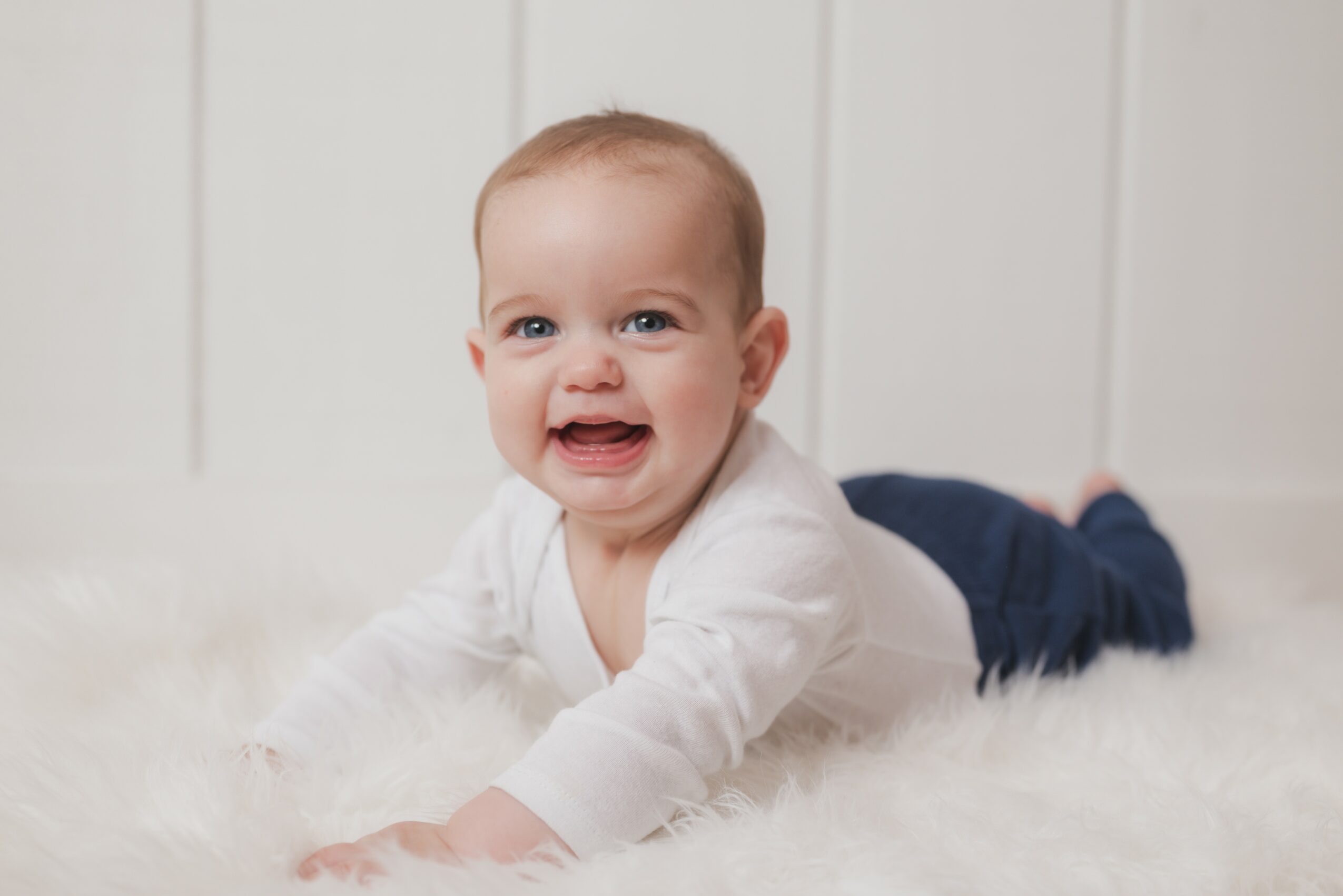 Baby lying on stomach on a soft surface, smiling and looking at the camera, with a white background.