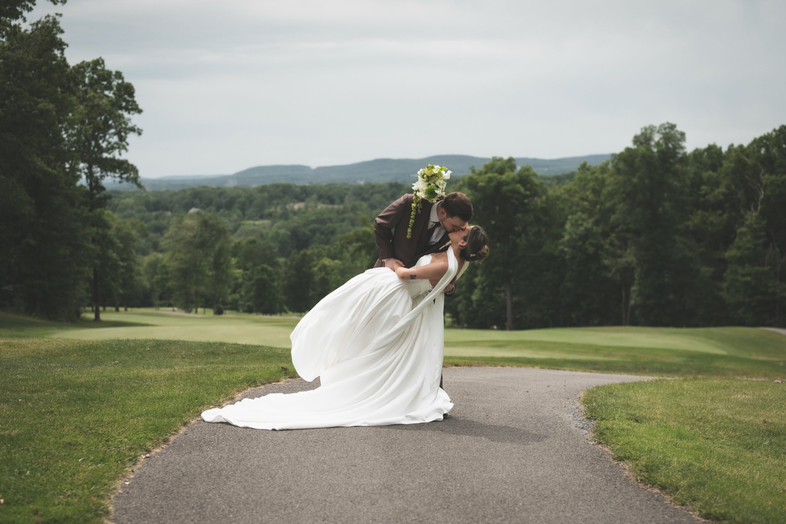 A bride in a white wedding dress holding a bouquet stands outdoors on a paved path with a landscape of trees and hills in the background.