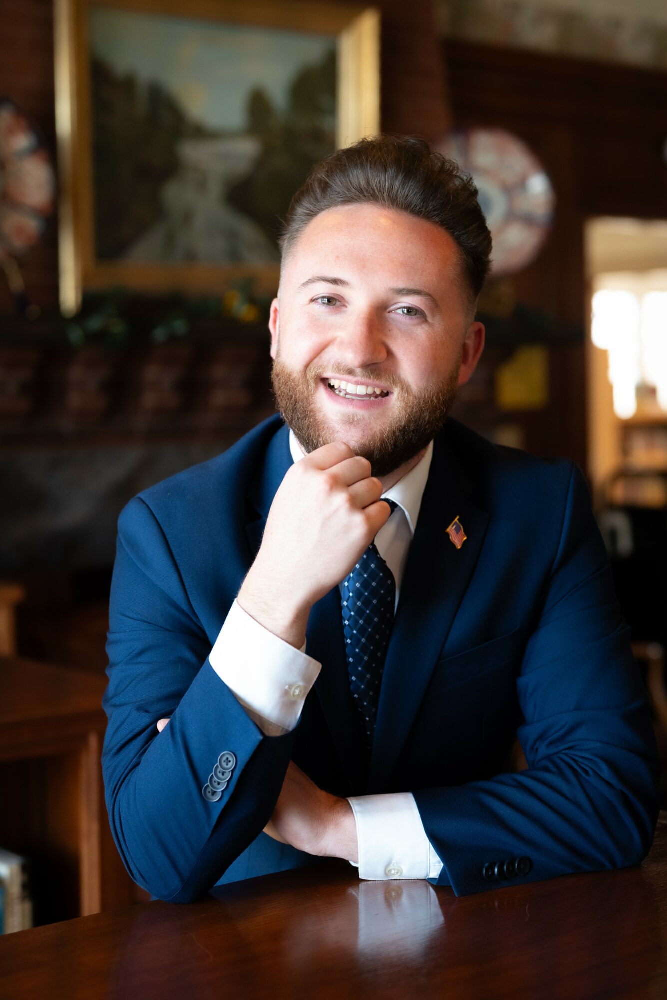 Smiling man in a blue suit with a white shirt and tie, sitting at a wooden table in a room with artwork and decor.