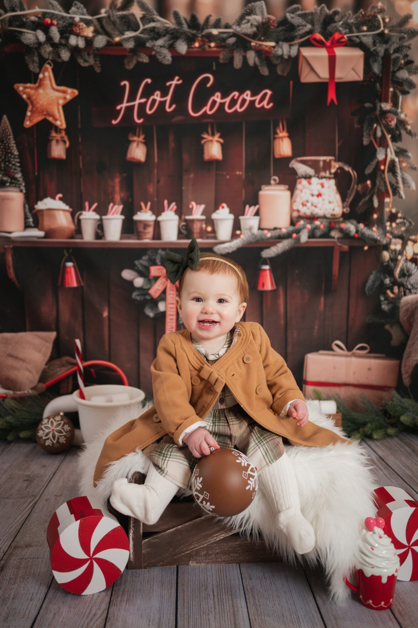 Child sitting on a wooden floor with holiday decorations, smiling, surrounded by a hot cocoa stand, candy cane, and festive items.