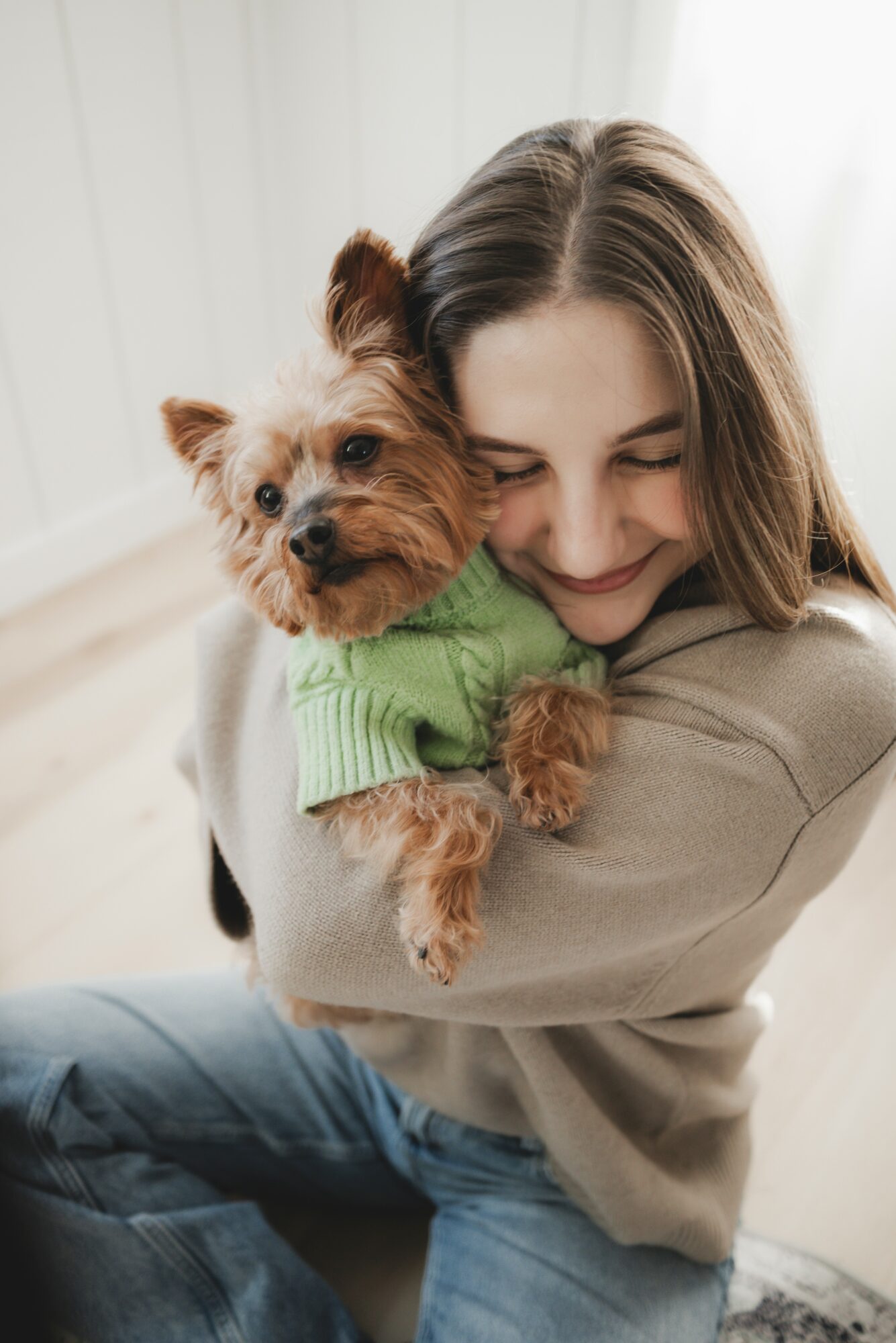 Young woman hugging a small dog wearing a green sweater, smiling with eyes closed, indoors with natural light.