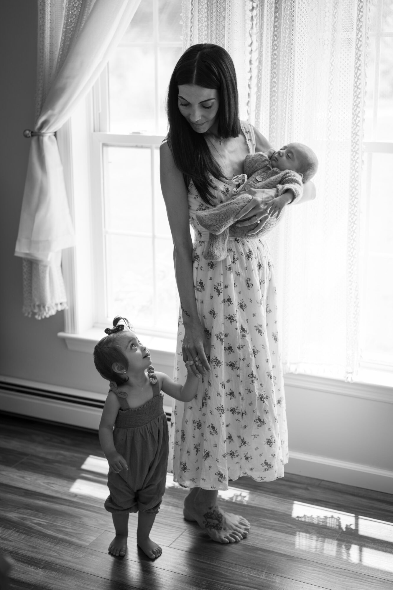 Woman holding a baby, young girl holding woman's hand, standing by a window with curtains, in a room with wooden floor.