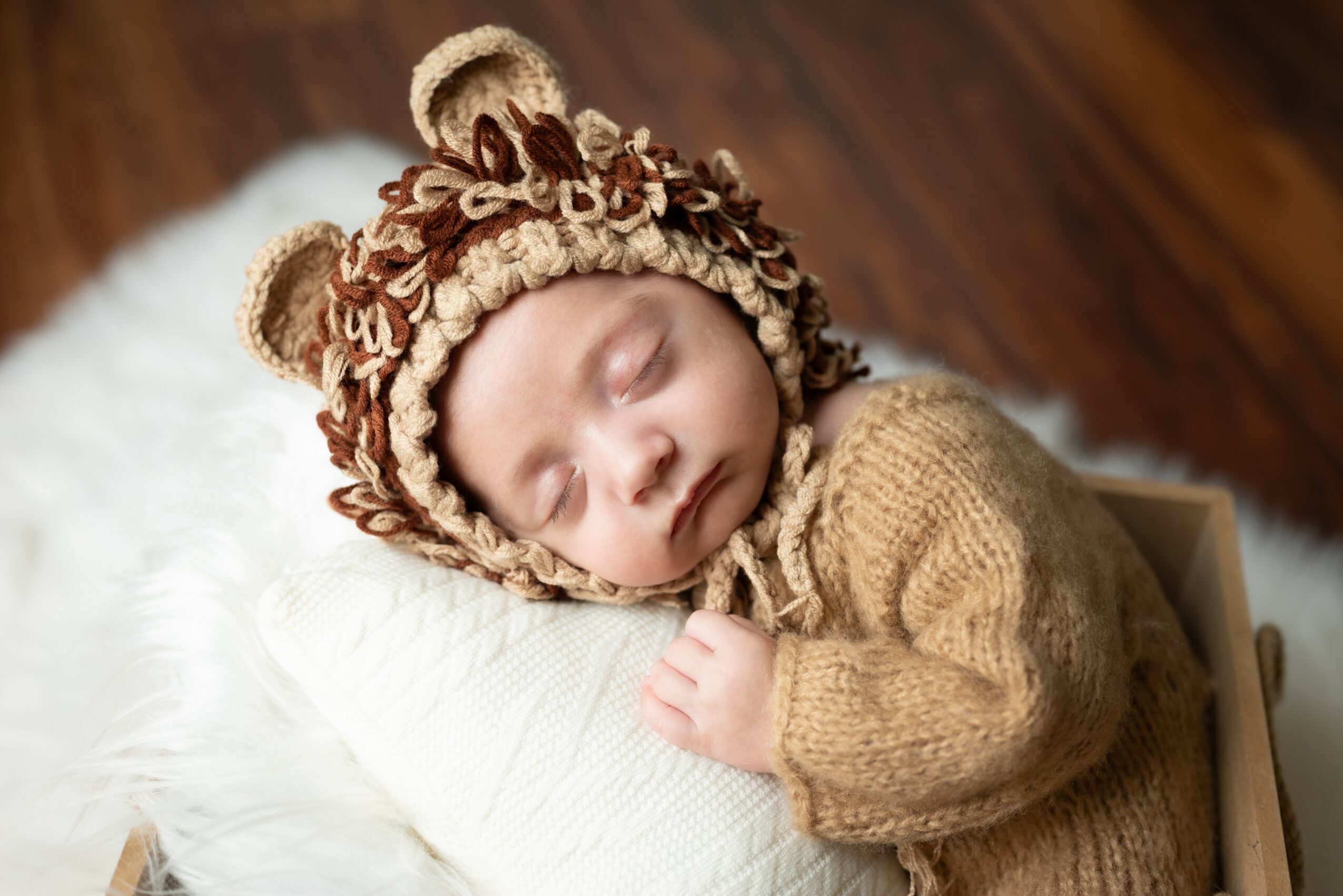 Baby sleeping on a white pillow, wearing a bear-themed hat and a brown sweater, lying on a soft surface.