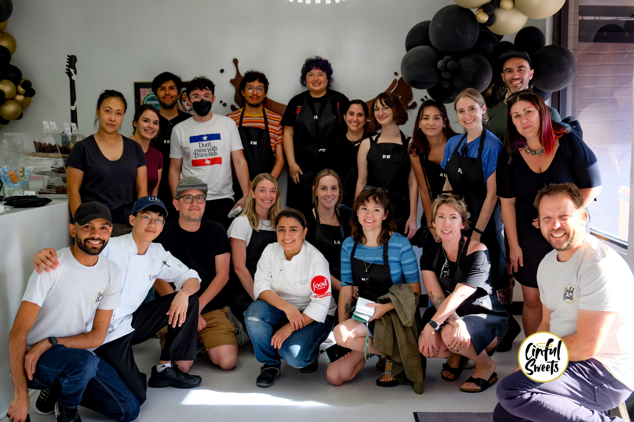 Group of people posing indoors with balloons and decorations, some smiling and some with neutral expressions.
