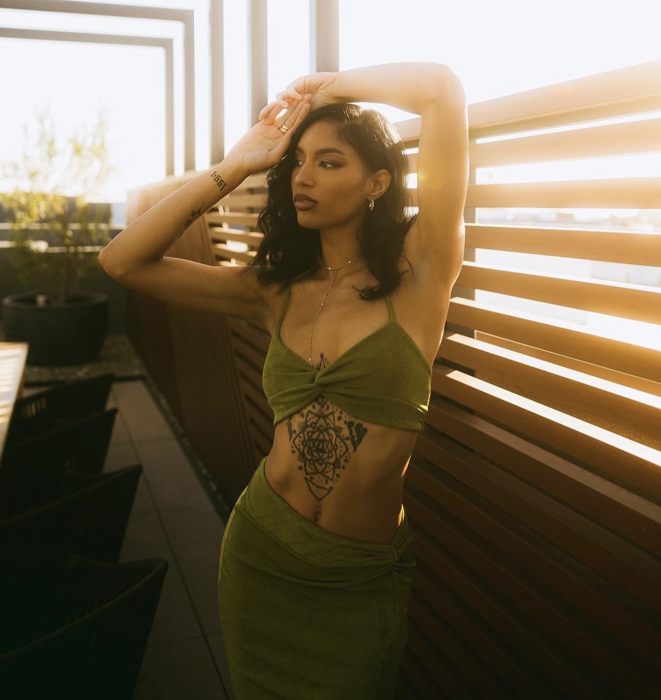 Woman with dark hair in a green crop top and skirt, standing outdoors near wooden slats, sunlight behind her.