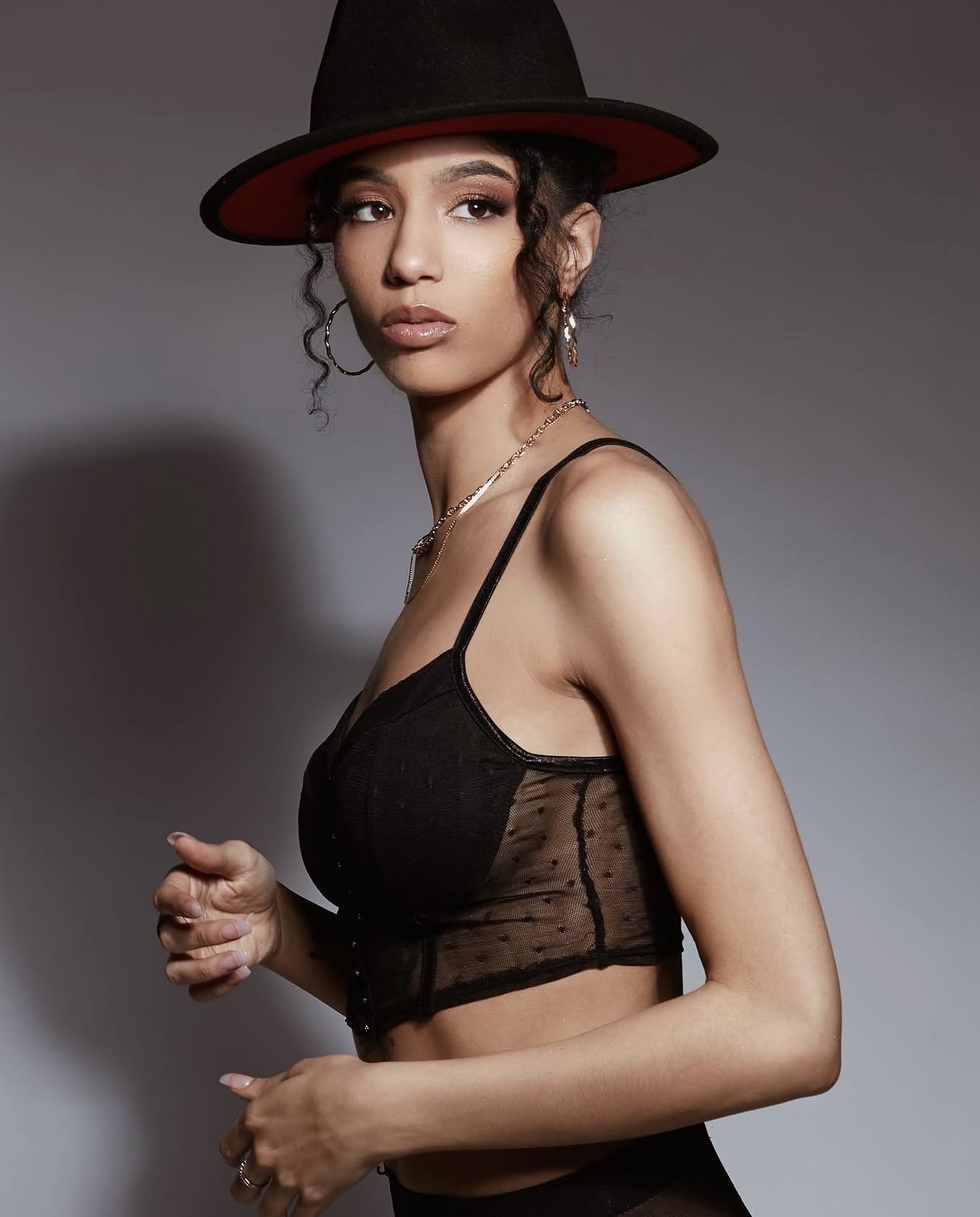 Young woman wearing a black sheer top and a wide-brimmed hat, looking at the camera with a neutral expression.