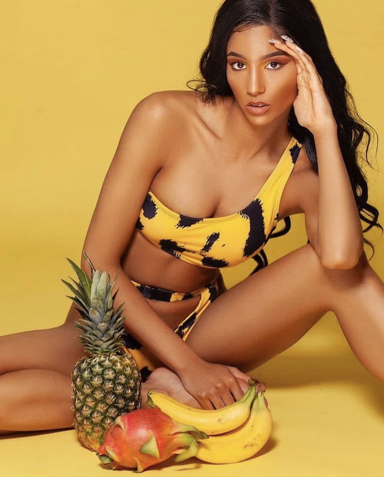 Woman with long dark hair in a yellow and black patterned outfit, sitting with fruits including pineapple, bananas, and apple, against yellow background.