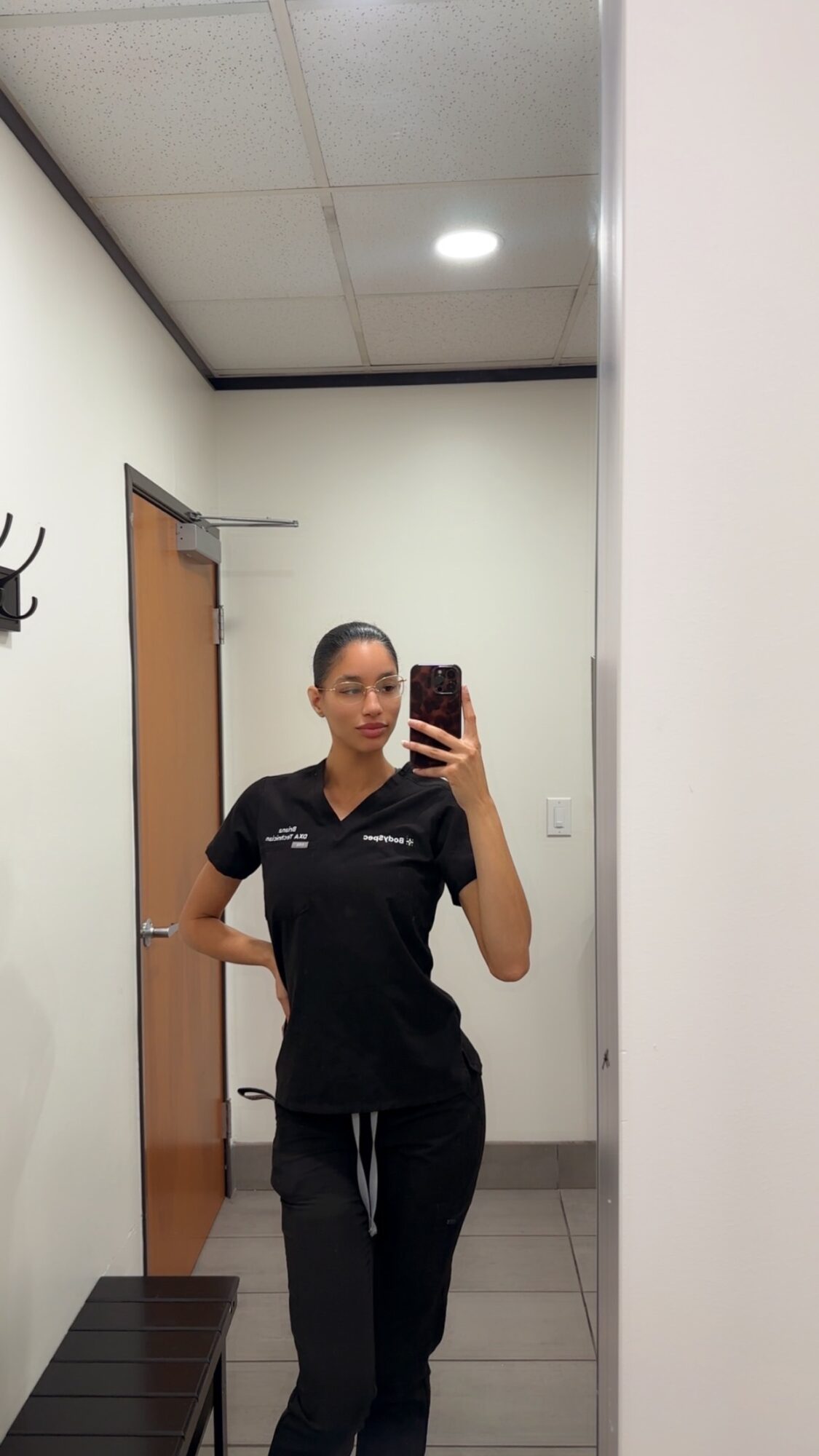 Woman in black uniform taking a mirror selfie in a room with white walls and a wooden door