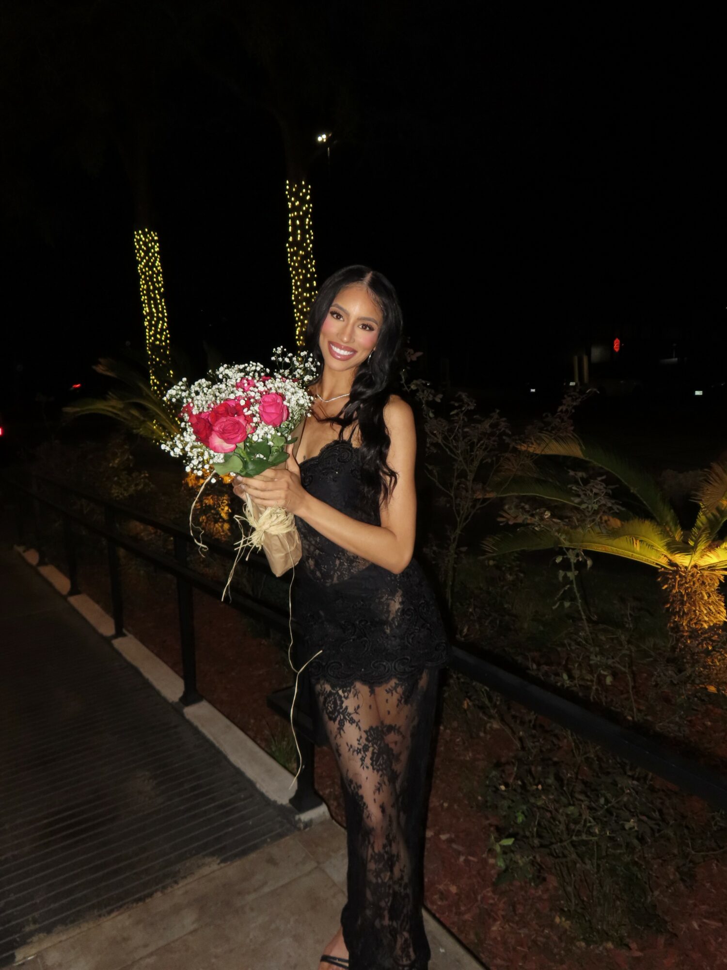 Woman in black lace dress holding a bouquet of flowers outdoors at night, smiling, with illuminated trees in background.