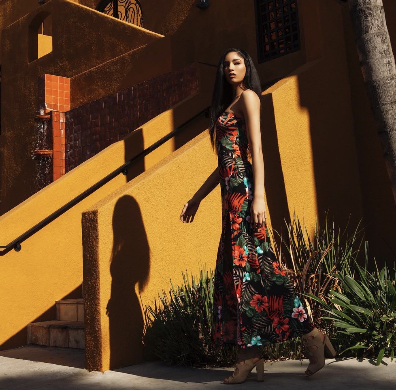Woman in a floral dress standing outdoors with a yellow wall and shadow, plants at her feet, sunlight casting shadows.