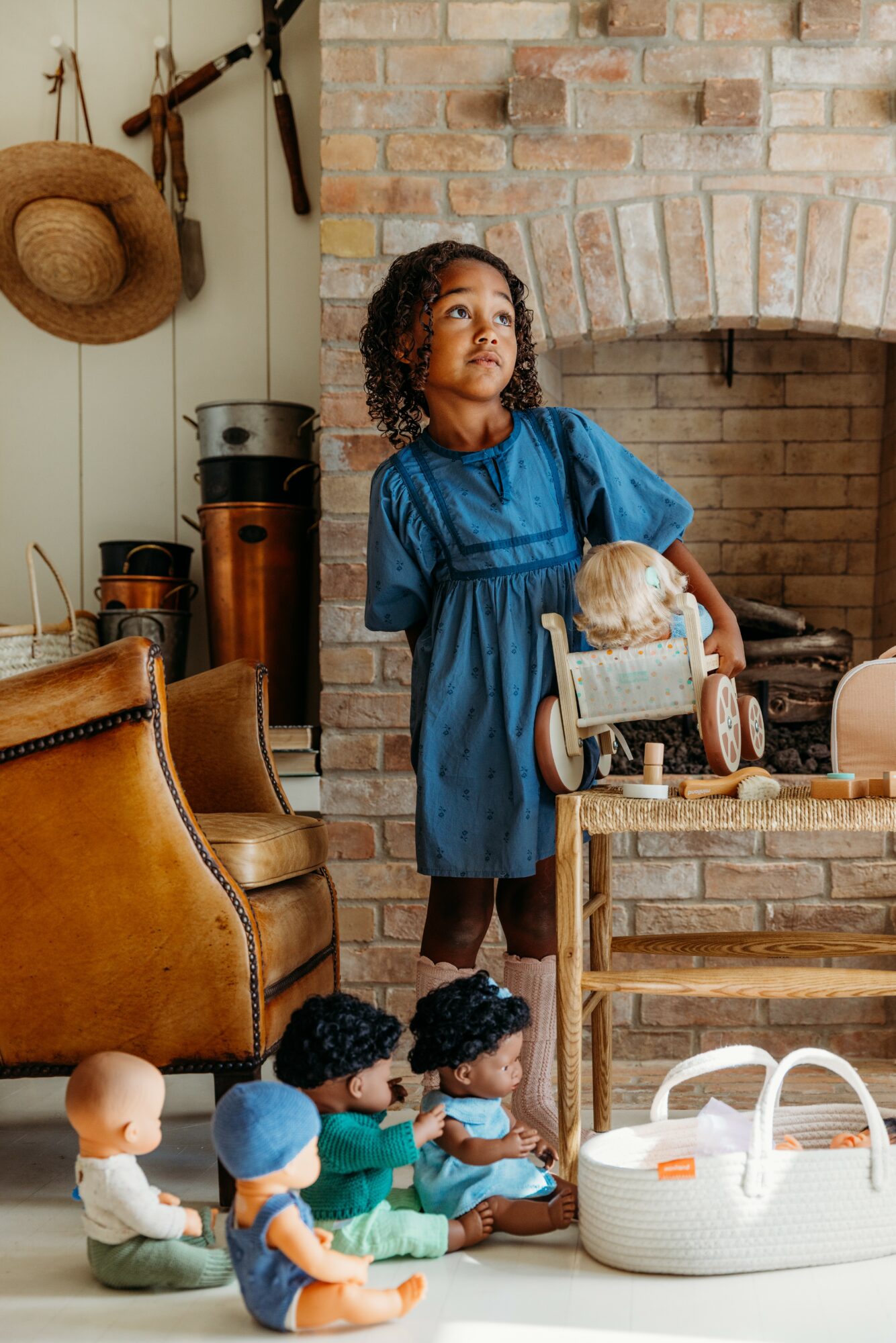 Young girl in blue dress standing near a fireplace with dolls and toys on the floor, cozy room setting.