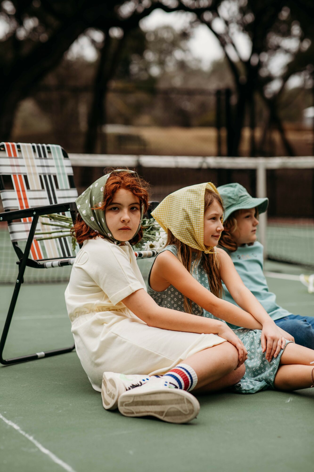 Three children sitting on a tennis court, two girls wearing hooded shirts and a boy in a cap, outdoors.