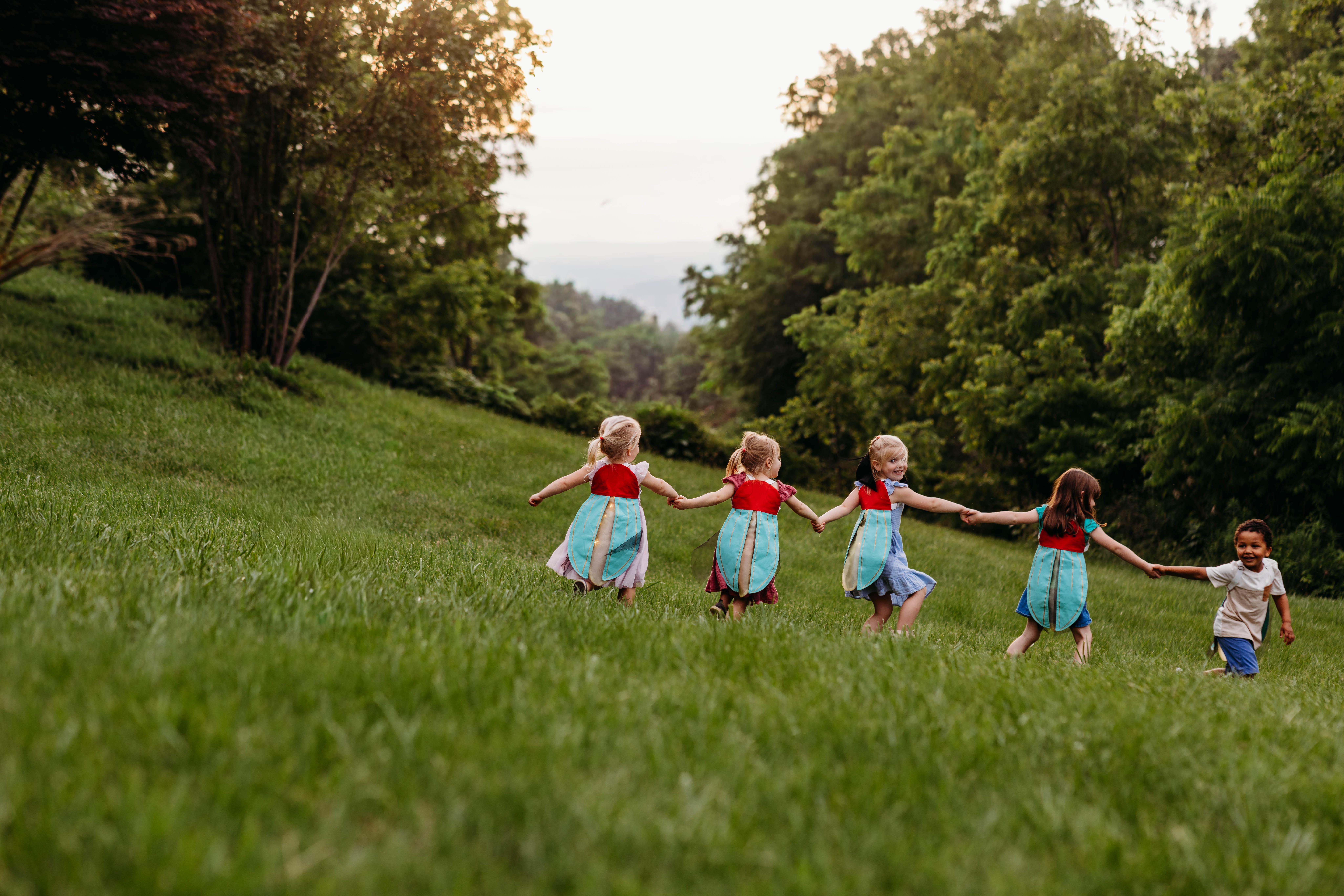 Five children holding hands and walking through a grassy field with trees in the background.