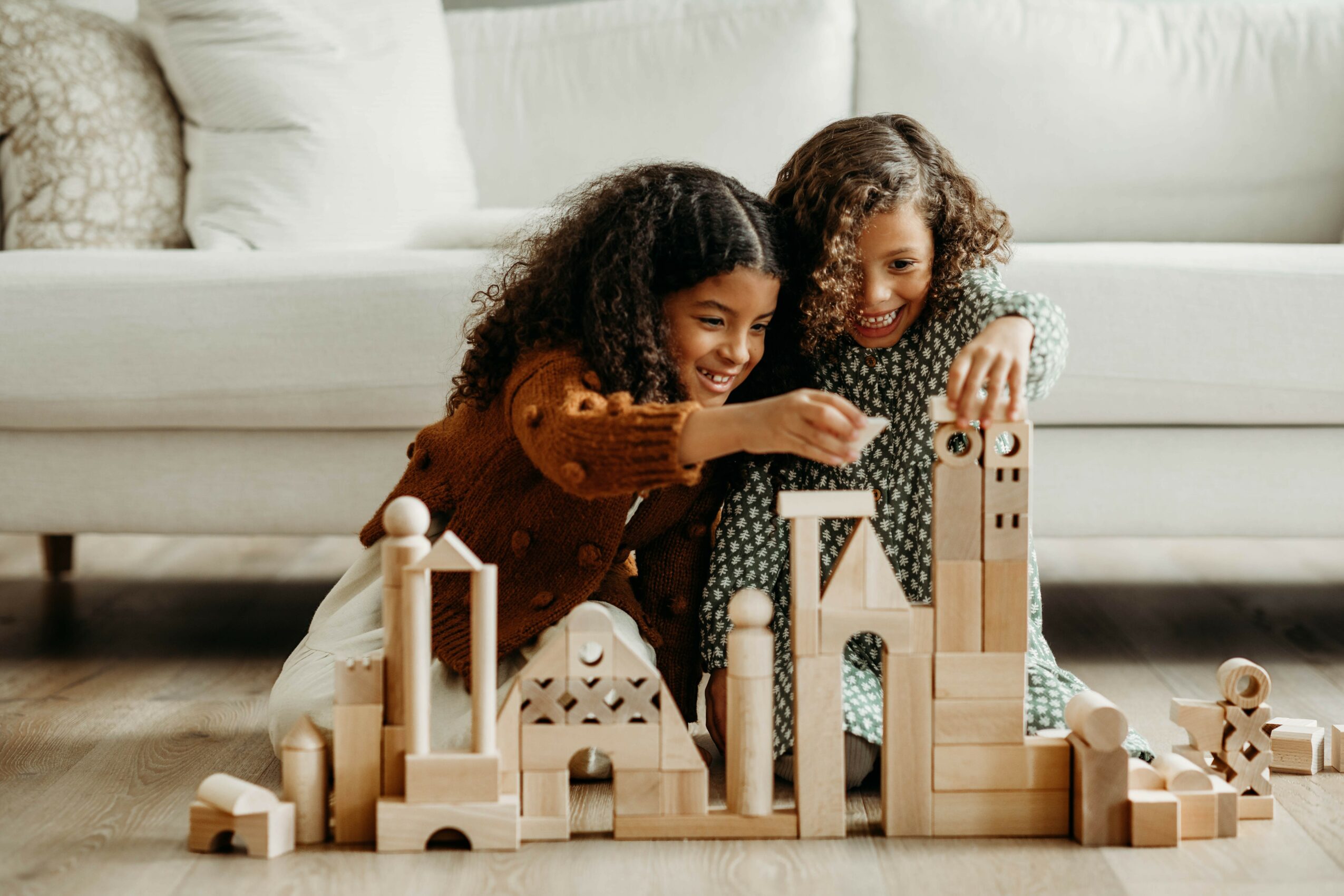 Two children with curly hair playing with wooden building blocks on the floor, smiling and engaging with the toys.