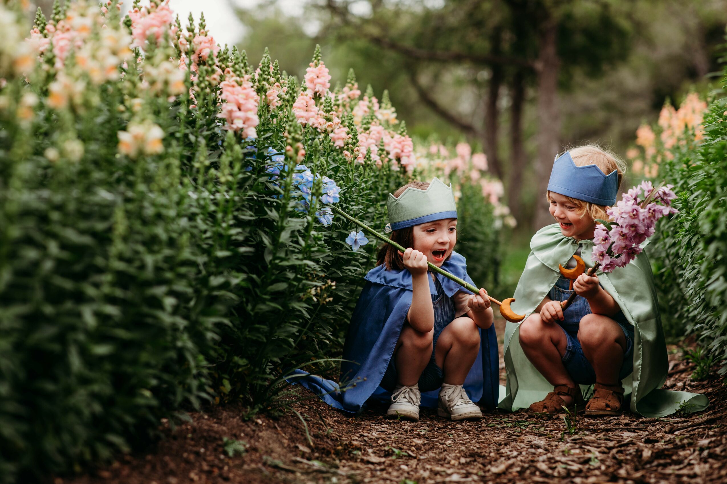 Two children wearing hats and capes sit on the ground among colorful flowers, playing with a stick and a flower bouquet.