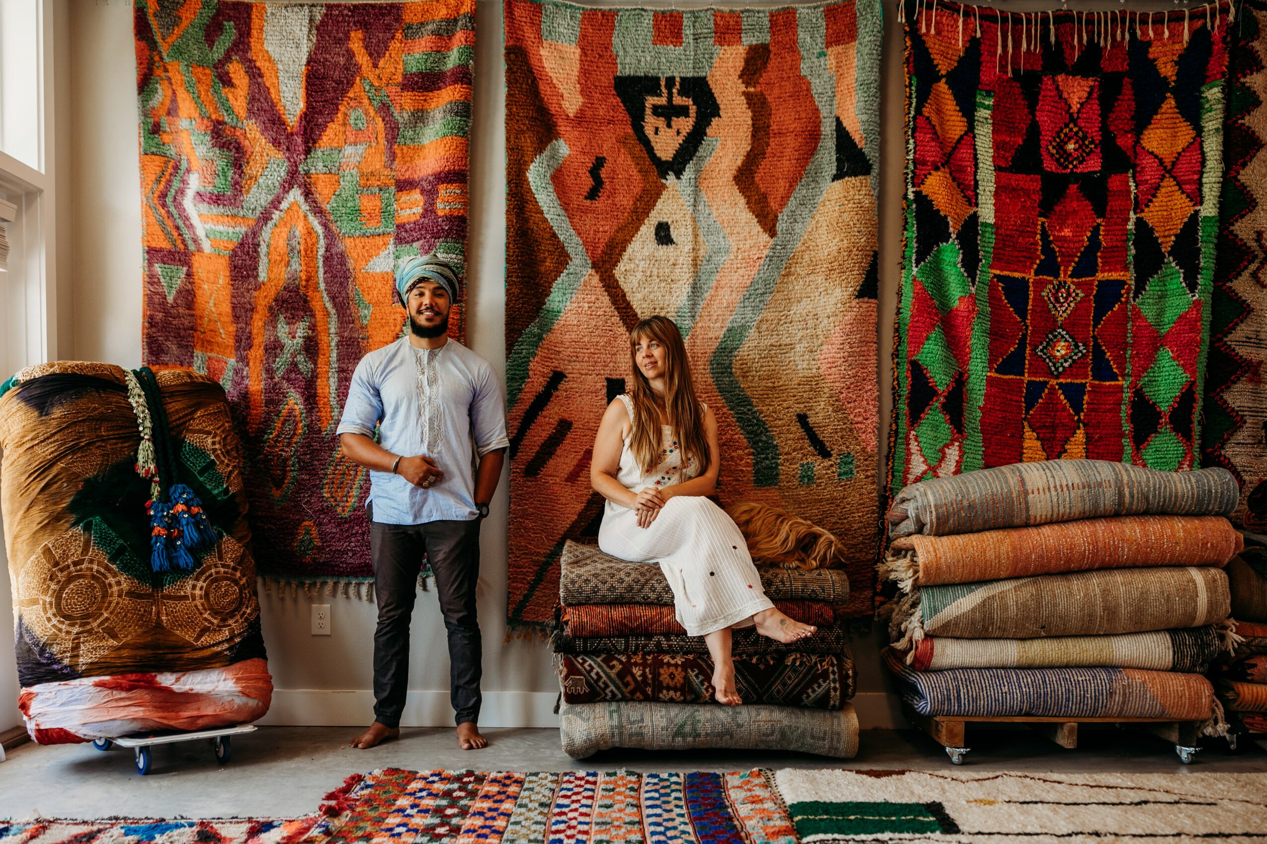 Two people in a room with colorful woven tapestries hanging on the wall, one sitting and one standing, with stacks of fabric nearby.