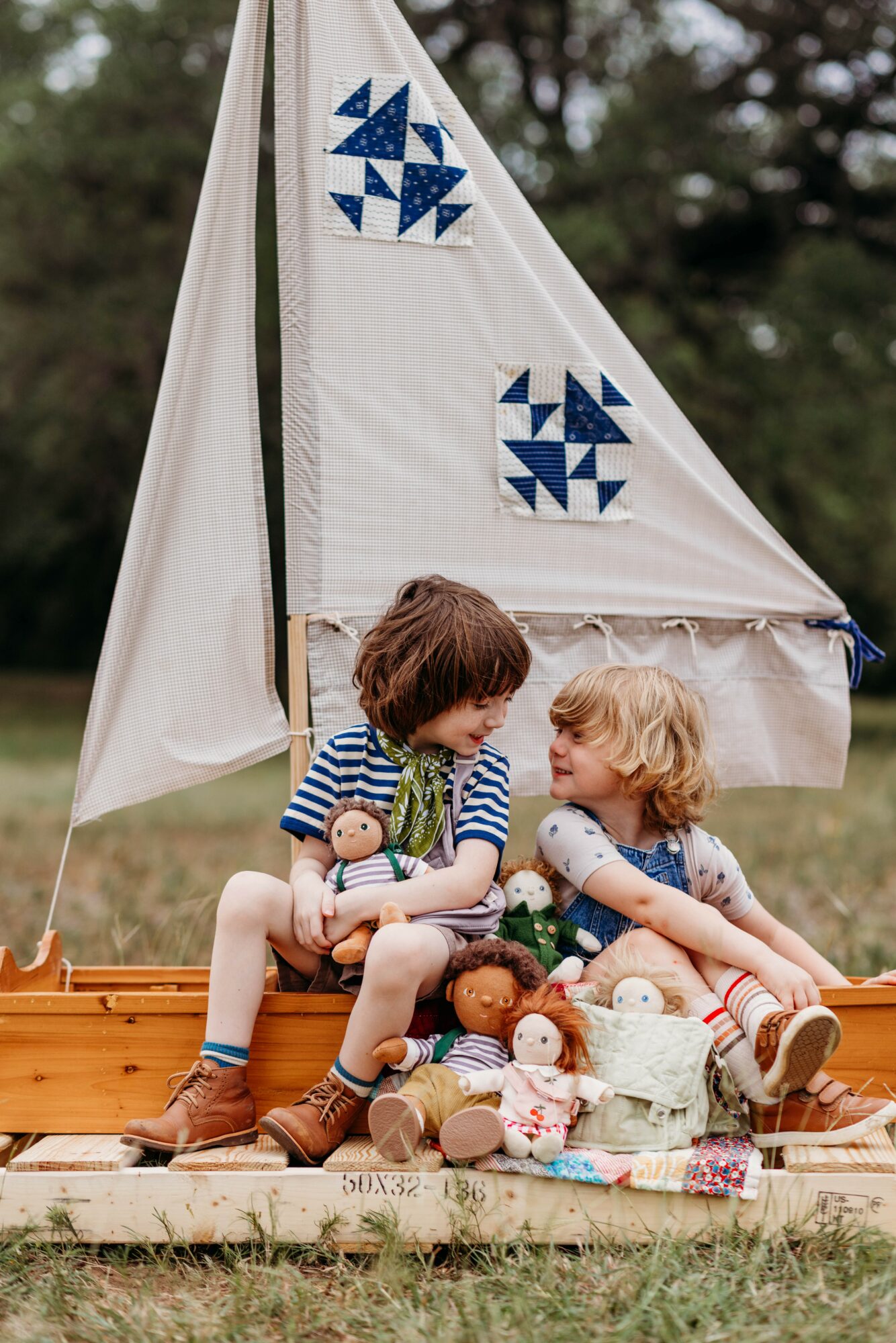 Two children sitting on a wooden boat with stuffed animals, in front of a small sailboat with blue and white sails, outdoors.