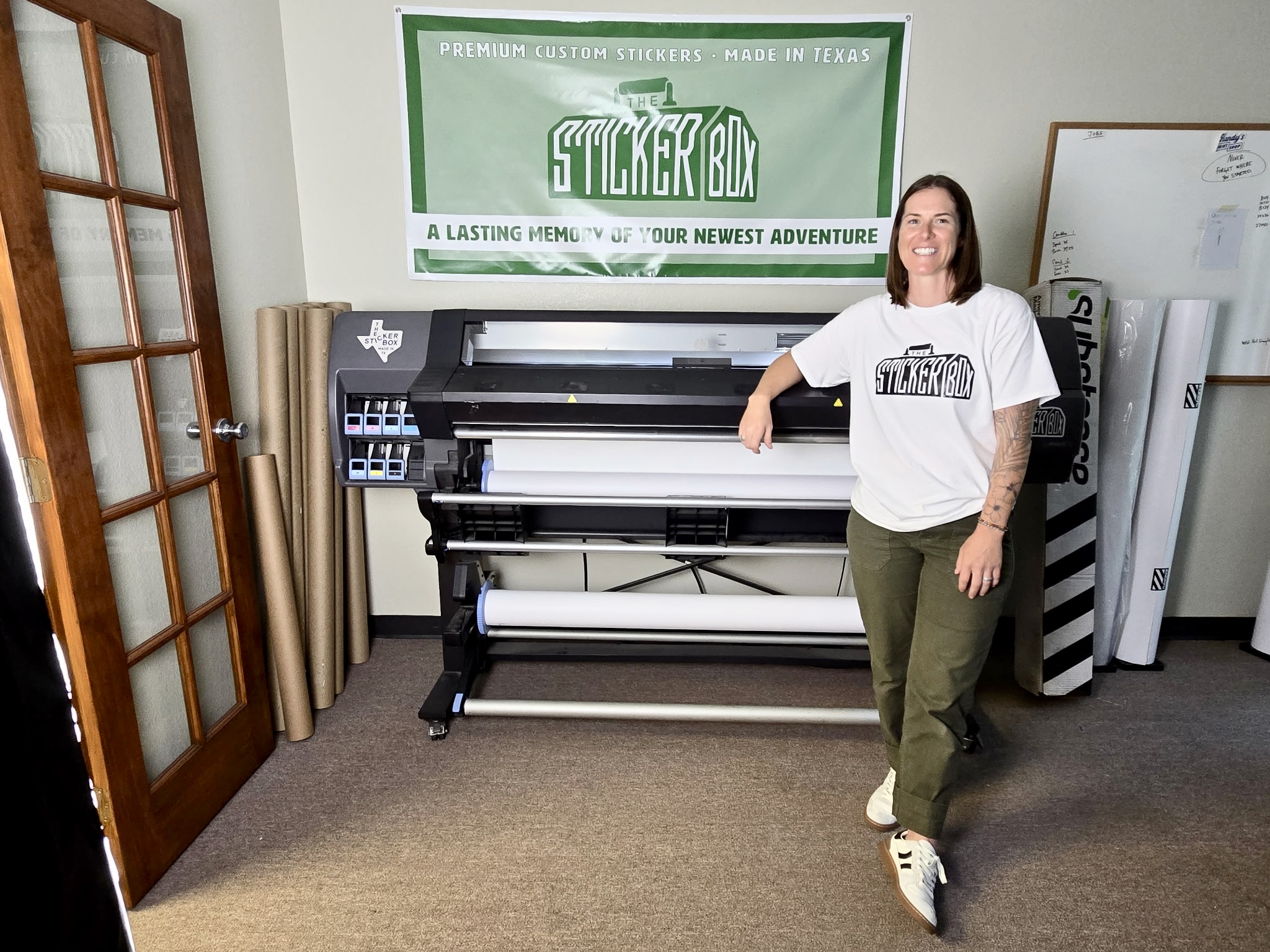 Woman standing next to a large printer in a room with a whiteboard and a poster on the wall.